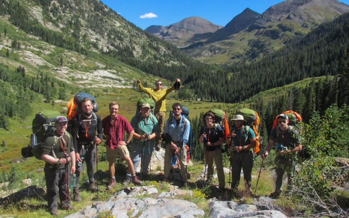 A group of people wearing backpacks pose for a photo in green mountainous valley.